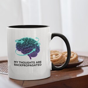 Könnte beinhalten: Weißer Keramikbecher mit schwarzem Innenraum und Griff. Der Becher zeigt eine türkis-violette Gehirngrafik mit Pfeilen und dem Text "MY THOUGHTS ARE BACKPROPAGATED". Der Becher steht auf einem Holztisch.
