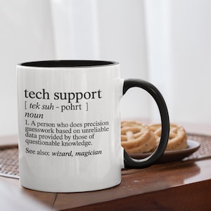 May include: White ceramic mug with a black interior and handle. The mug features the text "tech support" with a humorous definition, including the words "wizard" and "magician". The mug is on a wooden surface.