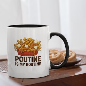 May include: White ceramic mug with a black interior and handle. The mug features a cartoon illustration of poutine, with the text "Poutine is my Routine". A plate of pretzels is in the background.