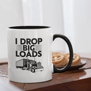 Peut inclure: Mug en céramique blanche avec intérieur et anse noirs. Le mug affiche le texte "I DROP BIG LOADS" au-dessus d'une illustration en noir et blanc d'un semi-remorque. Le mug est posé sur une table en bois.