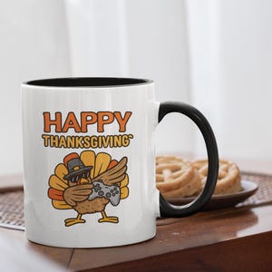 May include: White ceramic mug with a black interior and handle. The mug features a cartoon turkey wearing a hat and holding a game controller, with the words "HAPPY THANKSGIVING" in orange and yellow.