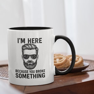Może przedstawiać: Biały ceramiczny kubek z czarnym wnętrzem i uchwytem. Kubek zawiera napis "I'M HERE BECAUSE YOU BROKE SOMETHING" oraz grafikę brodatego mężczyzny w okularach przeciwsłonecznych. Na tle talerz z ciastkami.
