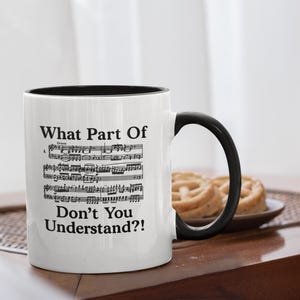 Puede incluir: Taza de cerámica blanca con interior y asa negros. La taza presenta el texto "What Part Of Don't You Understand?!" sobre una partitura musical. La taza está sobre una mesa de madera con un plato de pasteles en el fondo.