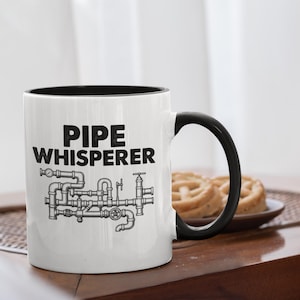 Könnte beinhalten: Weißer Keramikbecher mit schwarzem Innenraum und Henkel. Der Becher trägt die Aufschrift "PIPE WHISPERER" in fetten schwarzen Buchstaben über einer detaillierten Illustration von Rohren und Ventilen. Der Becher steht auf einer Holzoberfläche.