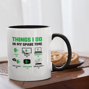 Op de afbeelding: Witte keramische mok met een zwarte binnenkant en handvat. De mok heeft de tekst "THINGS I DO IN MY SPARE TIME" in het groen, samen met iconen en tekst gerelateerd aan AI, zoals "PLAY AI", "WATCH AI" en "RESEARCH AI".