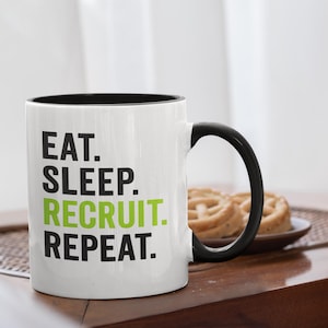 以下が含まれることがあります： 黒い内側とハンドルが付いた白いセラミックマグ。マグには「EAT. SLEEP. RECRUIT. REPEAT.」というテキストが黒と緑の文字で表示されています。マグは木製のテーブルの上にあり、背景には食べ物のプレートがあります。