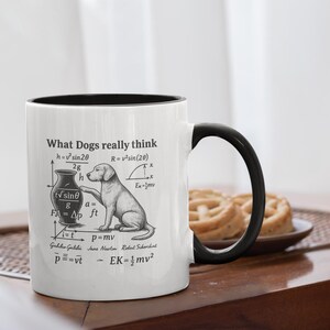 Könnte beinhalten: Weißer Keramikbecher mit schwarzem Innenraum und Henkel. Der Becher zeigt den Text "What Dogs really think" zusammen mit einem wissenschaftlichen Diagramm und einer Hundezeichnung. Der Becher steht auf einem Holztisch.