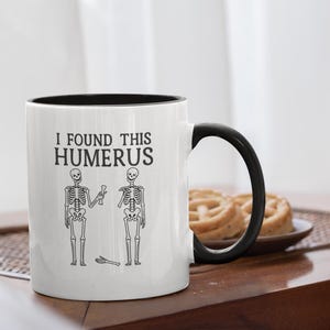 May include: White ceramic mug with a black interior and handle. The mug features the text "I FOUND THIS HUMERUS" with illustrations of skeletons. A humorous, medical-themed gift.