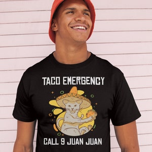 Pode incluir: Uma camiseta preta com um gr&aacute;fico branco de um gato usando um sombrero e segurando um taco. O texto "Taco Emergency" e "Call 9 Juan Juan" est&aacute; impresso na camiseta.