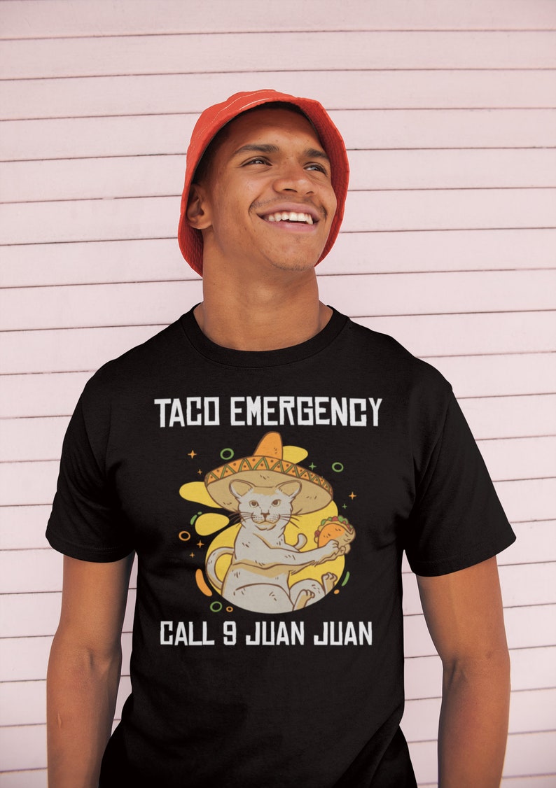 Pode incluir: Uma camiseta preta com um gr&aacute;fico branco de um gato usando um sombrero e segurando um taco. O texto "Taco Emergency" e "Call 9 Juan Juan" est&aacute; impresso na camiseta.