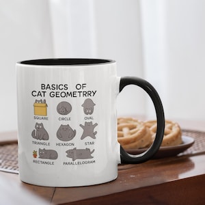 Puede incluir: Taza de cerámica blanca con interior y asa negros. La taza presenta el texto "BASICS OF CAT GEOMETRY" e ilustraciones de gatos en varias formas geométricas. La taza mide aproximadamente 10 cm de alto.