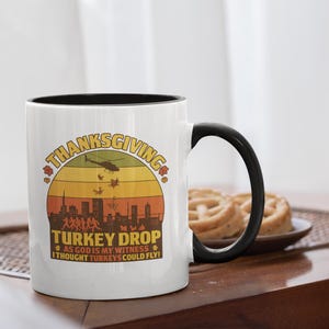 Op de afbeelding: Witte keramische mok met een zwarte handgreep en rand. De mok heeft een retro-stijl grafische afbeelding met de tekst "Thanksgiving Turkey Drop" en een silhouet van een stadsgezicht. De zin "As God is my witness, I thought turkeys could fly!" is ook opgenomen.
