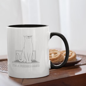 May include: White ceramic mug with a black interior and handle. The mug features a black and white illustration of a cat sitting on a laptop, with the text "I'M A PURRGRAMMER" below the laptop. The mug is on a wooden table.