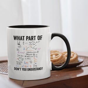 Könnte beinhalten: Weißer Keramikbecher mit schwarzem Innenraum und Henkel. Der Becher zeigt den Text "WHAT PART OF DON'T YOU UNDERSTAND?" zusammen mit mathematischen Gleichungen und Symbolen in Schwarz und verschiedenen Farben. Der Becher steht auf einem Holztisch.