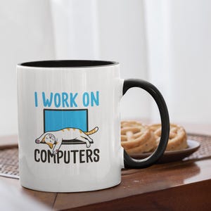 Pode incluir: Caneca de cerâmica branca com alça e borda pretas. A caneca apresenta o texto "I WORK ON COMPUTERS" com um desenho de gato. O gato está deitado numa tela de computador azul. No fundo, um prato com biscoitos.