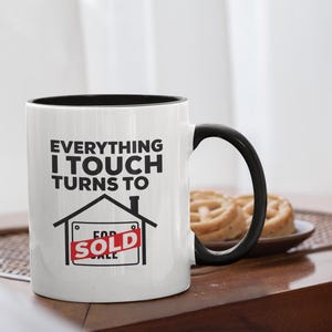 Puede incluir: Taza de cerámica blanca con interior y asa negros. La taza presenta el texto "EVERYTHING I TOUCH TURNS TO" sobre un gráfico de una casa con un letrero de "SOLD". La taza está sobre una superficie de madera.