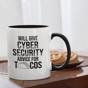 May include: White ceramic mug with a black interior and handle. The mug features the text "WILL GIVE CYBER SECURITY ADVICE FOR TACOS" in black, along with a taco illustration. The mug is on a wooden table.
