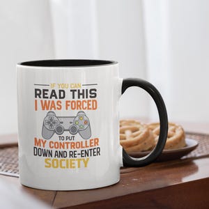 May include: White ceramic mug with a black interior and handle. The mug features text in black and orange that reads, "IF YOU CAN READ THIS I WAS FORCED TO PUT MY CONTROLLER DOWN AND RE-ENTER SOCIETY." A gaming-themed gift.