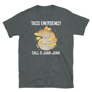 Pode incluir: Uma camiseta cinza escura mesclada com um gr&aacute;fico branco de um gato usando um sombrero e segurando um taco. O texto "Taco Emergency" est&aacute; acima do gato e "Call 9 Juan Juan" est&aacute; abaixo do gato.