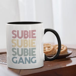 Op de afbeelding: Witte keramische mok met een zwarte handgreep en rand. De mok heeft de woorden "SUBIE SUBIE SUBIE GANG" in rode, gele en blauwe retro-letters. De mok staat op een houten tafel met een bord met gebak.
