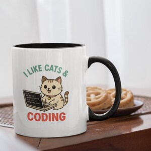 以下が含まれることがあります： 黒い内側とハンドルが付いた白いセラミックマグ。マグには「I LIKE CATS & CODING」というテキストと、ラップトップを使用している漫画の猫が描かれています。猫は明るい茶色で、濃い茶色の縞模様です。