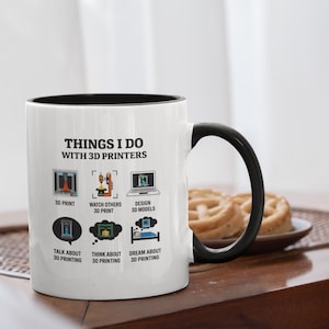 Könnte beinhalten: Weißer Keramikbecher mit schwarzem Innenraum und Henkel. Der Becher zeigt den Text "THINGS I DO WITH 3D PRINTERS" und Illustrationen von 3D-Druckaktivitäten. Ein Teller mit Backwaren steht daneben.