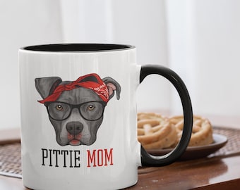 May include: White ceramic mug with a black handle and rim. The mug features a graphic of a pit bull wearing a red bandana and glasses, with the text "PITTIE MOM" in black and red.
