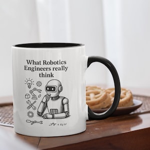 May include: White ceramic mug with a black interior and handle. The mug features the text "What Robotics Engineers really think" and a graphic of a robot and engineering tools. A plate of pastries is in the background.