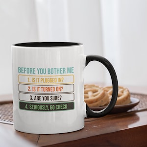 Peut inclure: Mug en céramique blanche avec intérieur et anse noirs. Le mug affiche le texte « BEFORE YOU BOTHER ME » suivi d'une liste numérotée : « 1. EST-CE BRANCHÉ ? » « 2. EST-CE ALLUMÉ ? » « 3. ÊTES-VOUS SÛR ? » « 4. SÉRIEUSEMENT, ALLEZ VÉRIFIER ».