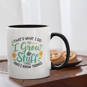 May include: White ceramic mug with a black interior and handle. The mug features the text "THAT'S WHAT I DO I GROW STUFF AND I KNOW THINGS" in green and orange lettering, along with gardening-themed illustrations. The mug is on a wooden table.