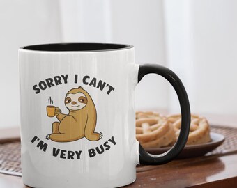 May include: White ceramic mug with a black interior and handle. The mug features a cartoon sloth holding a cup of coffee, with the text "SORRY I CAN'T I'M VERY BUSY". A plate of biscuits sits in the background.