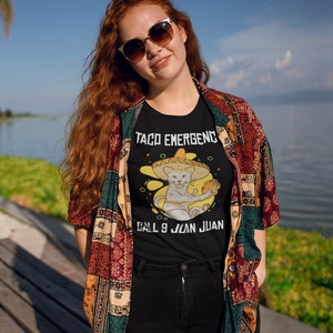 Pode incluir: Uma camiseta preta com um gato branco usando um sombrero e segurando um taco. O texto na camiseta diz "Taco EmergencY Call 9 Juan Juan".
