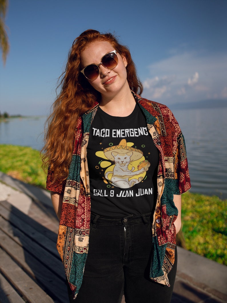 Pode incluir: Uma camiseta preta com um gato branco usando um sombrero e segurando um taco. O texto na camiseta diz "Taco EmergencY Call 9 Juan Juan".