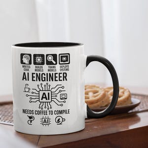 May include: White ceramic mug with a black interior and handle. The mug features the text "AI ENGINEER" and related icons, including "WRITES CODE," "BUILDS MODELS," and "NEEDS COFFEE TO COMPILE." A plate of biscuits is in the background.