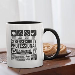 Könnte beinhalten: Weißer Keramikbecher mit schwarzem Innenraum und Henkel. Der Becher zeigt Text und Symbole zum Thema Cybersicherheit, darunter "Cybersecurity Professional" und "Warning: Sarcasm Inside."