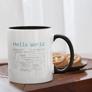 May include: White ceramic mug with a black interior and handle. The mug features the text "Hello World" in light blue, along with code snippets. The mug is on a wooden table, with a plate of pastries in the background.