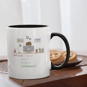 Pode incluir: Caneca de café branca com interior e alça pretos. A caneca apresenta um gráfico de uma configuração de computador com o texto "while !dead: eat() code() //sleep()". Um prato de biscoitos está por perto.