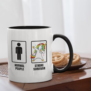 Può includere: Tazza in ceramica bianca con interno e manico neri. La tazza presenta due immagini incorniciate: un omino stilizzato con l'etichetta "NORMAL PEOPLE" e un unicorno che fa il dab, con l'etichetta "STROKE SURVIVOR". Un piatto di pretzel è sullo sfondo.