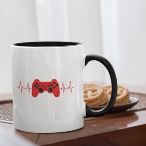 Puede incluir: Taza de cerámica blanca con interior y asa negros. Un gráfico de mando rojo está centrado en la taza, con una línea en forma de latido del corazón que se extiende desde el mando. La taza está sobre una mesa de madera.