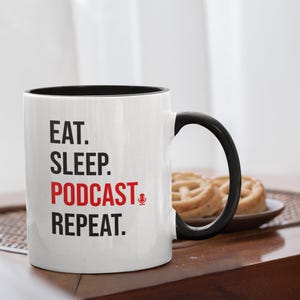 May include: White ceramic mug with a black interior and handle. The mug features the text "EAT. SLEEP. PODCAST. REPEAT." in black and red lettering, with a microphone graphic. The mug is on a wooden table.
