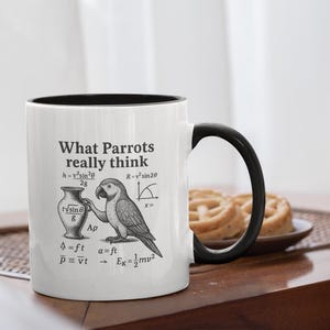 May include: White ceramic mug with a black interior and handle. The mug features the text "What Parrots really think" along with scientific formulas and a detailed illustration of a parrot. A plate of pastries sits in the background.