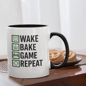May include: White ceramic mug with a black interior and handle. The mug features the words "WAKE BAKE GAME REPEAT" in black, with green icons. The mug is on a wooden table, with a plate of pastries in the background.