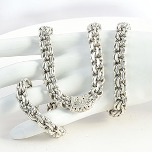 May include: A silver necklace and bracelet set with a chunky, woven chain design. The necklace has a decorative clasp with an ornate floral pattern. The jewellery is displayed on a white surface.
