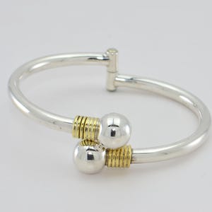Peut inclure: Un bracelet jonc en argent avec deux boules argentées et des accents dorés. Le bracelet est ouvert et possède une charnière.