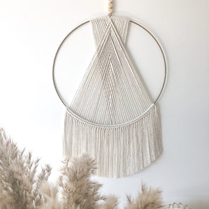 Peut inclure: Une suspension murale en macramé blanche avec un motif géométrique, comprenant un cercle en or et une frange de fil blanc.
