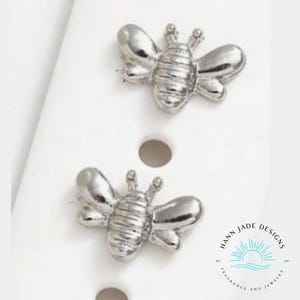 May include: A pair of silver bee stud earrings. The bees have a textured body and are detailed with wings and antennae.