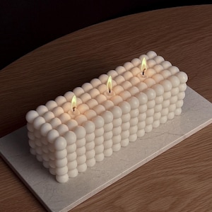 Könnte beinhalten: Eine weiße Kerze mit drei Dochten, aus kleinen runden Perlen gefertigt, steht auf einer weißen Marmorfläche. Die Kerze ist angezündet und die Flammen sind sichtbar.