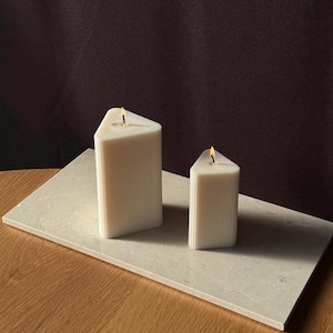 Könnte beinhalten: Zwei weiße dreieckige Kerzen mit einer gerippten Textur auf einer hellbeigen Oberfläche. Die Kerzen sind angezündet und haben eine kleine Flamme.