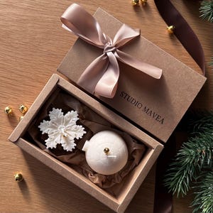 Op de afbeelding: Een lichtbruine geschenkdoos met een bijpassend lint. Binnenin liggen een witte sneeuwvlok en een bolvormig ornament op bruin papier. De doos is gemarkeerd met "STUDIO MAVEA".