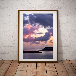 May include: A framed photograph depicting a dramatic sunset over a body of water. The sky is a mix of pink, purple, and grey clouds, with a sliver of orange light peeking through. The water is calm and reflective, with a small island in the distance.